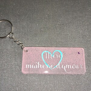 Peut inclure: Un porte-cl&eacute;s rectangulaire rose avec une cha&icirc;ne et un anneau argent&eacute;s. Le porte-cl&eacute;s porte les mots "Merci ma&icirc;tresse d'amour" en &eacute;criture blanche avec un c&oelig;ur turquoise. Une petite gemme violette est sertie sur le c&ocirc;t&eacute; droit.