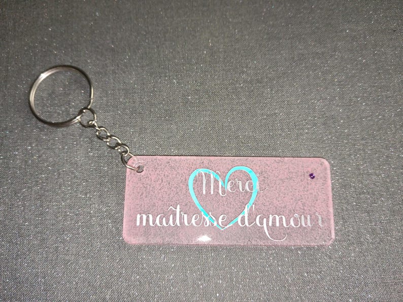 Peut inclure: Un porte-cl&eacute;s rectangulaire rose avec une cha&icirc;ne et un anneau argent&eacute;s. Le porte-cl&eacute;s porte les mots "Merci ma&icirc;tresse d'amour" en &eacute;criture blanche avec un c&oelig;ur turquoise. Une petite gemme violette est sertie sur le c&ocirc;t&eacute; droit.