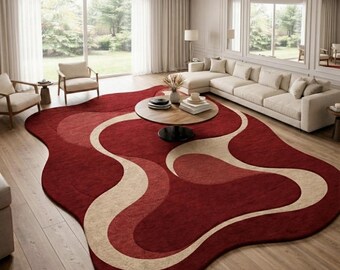May include: A large, abstract-shaped area rug in a living room setting. The rug features a deep red base with flowing, lighter red and beige wave-like patterns. The rug is placed on a hardwood floor, with a coffee table and sofa visible.