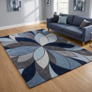 May include: A large area rug with a floral design in shades of blue, gray, and white. The rug features a central flower-like pattern with petal shapes radiating outwards. The rug is placed on a wooden floor in a living room setting.