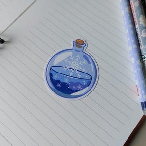 May include: A circular sticker showing a blue potion bottle with a cork. The bottle contains a snowflake design. The sticker is on a lined notebook with two pens.