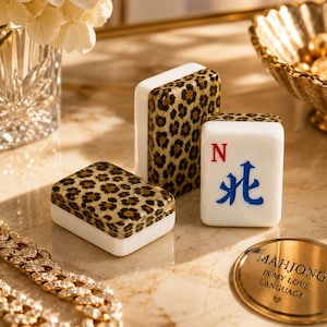 May include: Three white mahjong tiles featuring a brown and black leopard print pattern on the top surfaces. One tile displays the red letter N and a blue Chinese character. A gold circular token reads MAHJONG IS MY LOVE LANGUAGE.