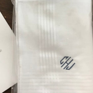 May include: A white handkerchief with a blue monogram of the letters "GHJ" in a circle. The handkerchief is folded and packaged in a clear plastic bag.