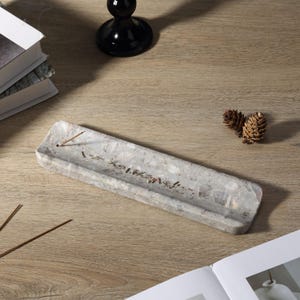 May include: A rectangular grey incense holder with a speckled pattern and text. The holder is made of stone and has a groove to catch ash. Incense sticks and pine cones are also visible on a wooden surface.