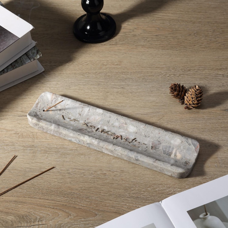 May include: A rectangular grey incense holder with a speckled pattern and text. The holder is made of stone and has a groove to catch ash. Incense sticks and pine cones are also visible on a wooden surface.