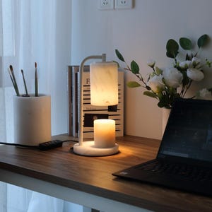 Op de afbeelding: Een kaarsverwarmerlamp van wit marmer met een cilindrische kap en een ronde voet. De lamp heeft een gebogen arm en straalt een warme, zachte gloed uit op een brandende kaars die direct onder de kap staat, wat zorgt voor een functioneel verlichtingsstuk.