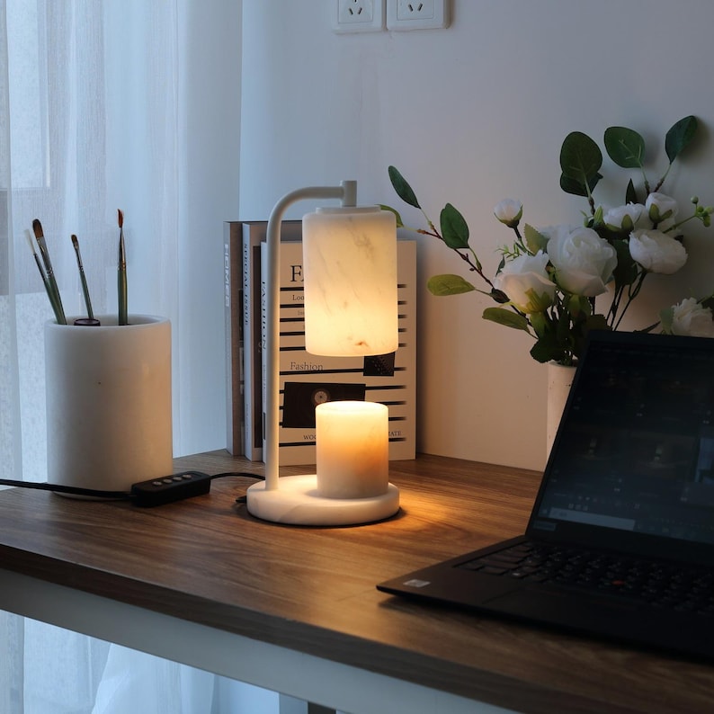 Op de afbeelding: Een kaarsverwarmerlamp van wit marmer met een cilindrische kap en een ronde voet. De lamp heeft een gebogen arm en straalt een warme, zachte gloed uit op een brandende kaars die direct onder de kap staat, wat zorgt voor een functioneel verlichtingsstuk.