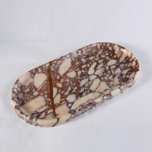 May include: An oval-shaped decorative tray with a marble pattern. The tray features a mix of brown, cream, and white colors, creating a natural stone appearance. The edges are slightly raised, and the overall design is smooth and polished.