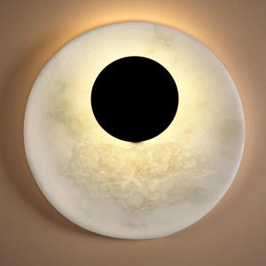 May include: A round, off-white wall sconce with a large, central black circle. The sconce has a marbled texture and emits a soft, warm light around the black circle. The fixture is designed for indoor use.