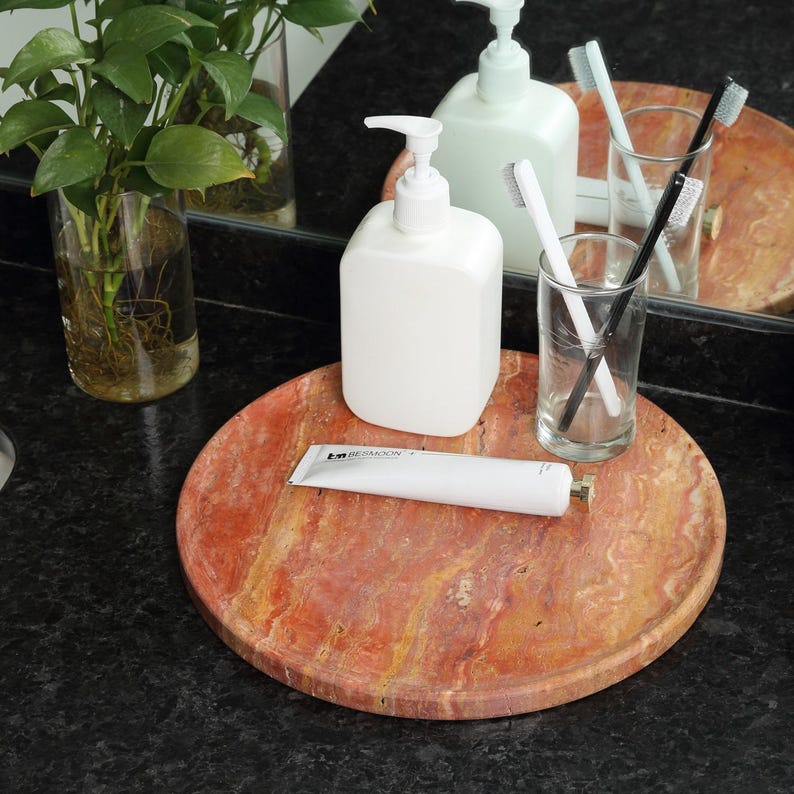 Puede incluir: Una bandeja redonda de m&aacute;rmol rojo-naranja sostiene un dispensador de jab&oacute;n blanco, un tubo de pasta de dientes y dos vasos transparentes con cepillos de dientes. Una planta verde en un jarr&oacute;n de cristal est&aacute; en el fondo. Los art&iacute;culos est&aacute;n sobre una encimera negra.