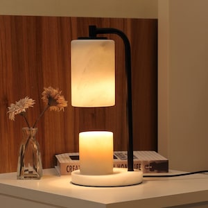 Op de afbeelding: Een zwarte metalen lamp met een cilindrische witte marmeren kap en een bijpassende witte marmeren voet. De lamp heeft een naar beneden gerichte lichtbron die een witte cilindrische kaars op de ronde marmeren voet verlicht.