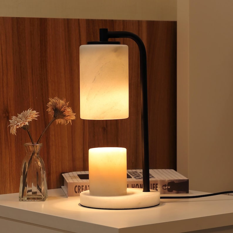 Op de afbeelding: Een zwarte metalen lamp met een cilindrische witte marmeren kap en een bijpassende witte marmeren voet. De lamp heeft een naar beneden gerichte lichtbron die een witte cilindrische kaars op de ronde marmeren voet verlicht.