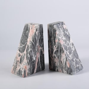 Puede incluir: Dos sujetalibros de mármol con forma triangular. Los sujetalibros son grises con vetas rosas y blancas. Los sujetalibros están en ángulo y se mantienen en posición vertical. Los sujetalibros son de piedra y probablemente pesados.