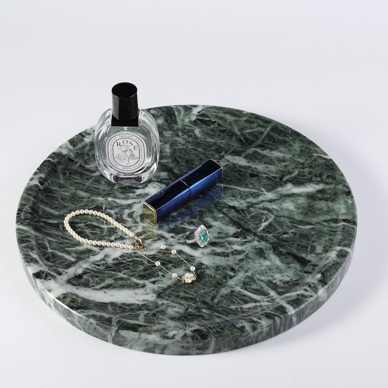Puede incluir: Una bandeja redonda de m&aacute;rmol verde con un frasco de perfume, l&aacute;piz labial azul, un collar de perlas y un anillo con una piedra verde. La bandeja tiene un patr&oacute;n veteado natural, ideal para exhibir joyas y productos de belleza.