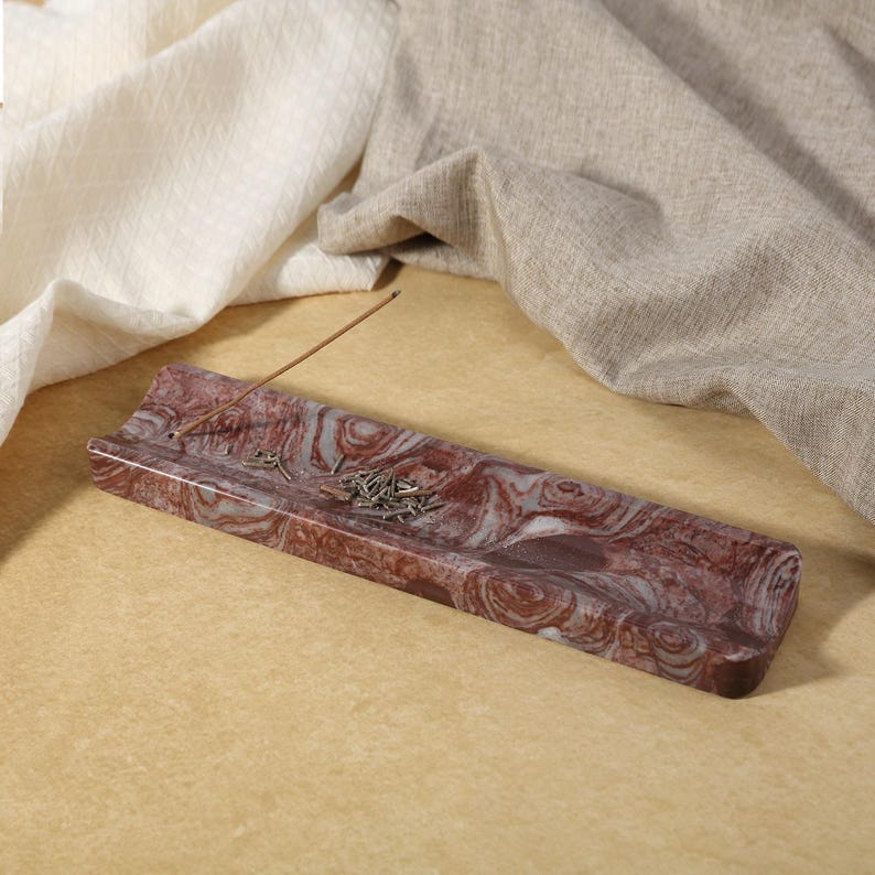 May include: A rectangular incense holder made of red and white swirled stone. An incense stick is placed in the holder, with a small pile of incense. The holder is on a light brown surface, with beige and white fabric in the background.
