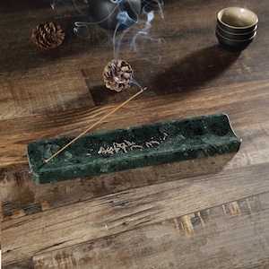 May include: A dark green marble incense holder, rectangular in shape. Incense is burning, releasing smoke. The holder sits on a wooden surface, with pine cones and small bowls in the background.