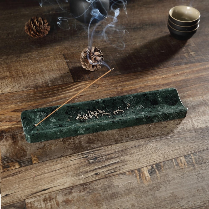 May include: A dark green marble incense holder, rectangular in shape. Incense is burning, releasing smoke. The holder sits on a wooden surface, with pine cones and small bowls in the background.