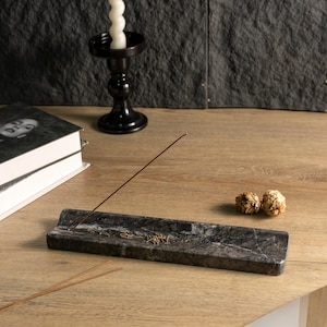 May include: A dark grey marble incense holder with a brown incense stick. The holder sits on a light wood surface, with a black candle holder and books in the background. Two small, dried decorative items are also present.