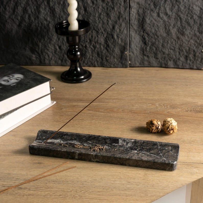 May include: A dark grey marble incense holder with a brown incense stick. The holder sits on a light wood surface, with a black candle holder and books in the background. Two small, dried decorative items are also present.
