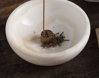 May include: A white and beige marble incense holder with a brass incense stick holder. The incense stick is lit and emitting smoke. The holder is filled with incense ash and small pieces of incense.