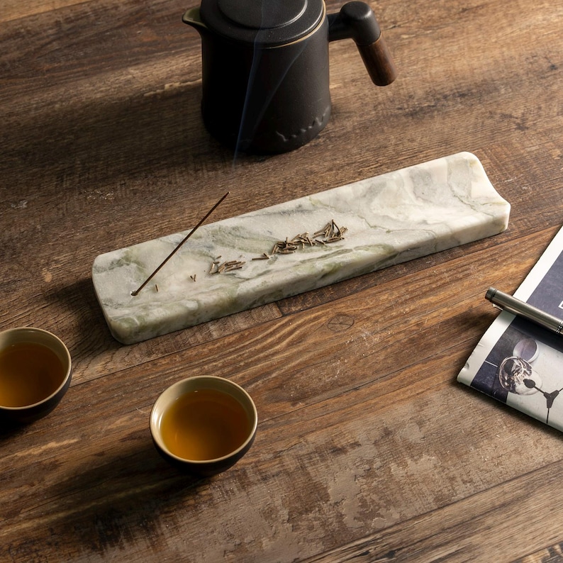 May include: A rectangular marble incense holder with a green and white pattern, holding an incense stick and loose incense. A black teapot with a wooden handle is above the holder. Two small teacups with tea are on a wooden surface.