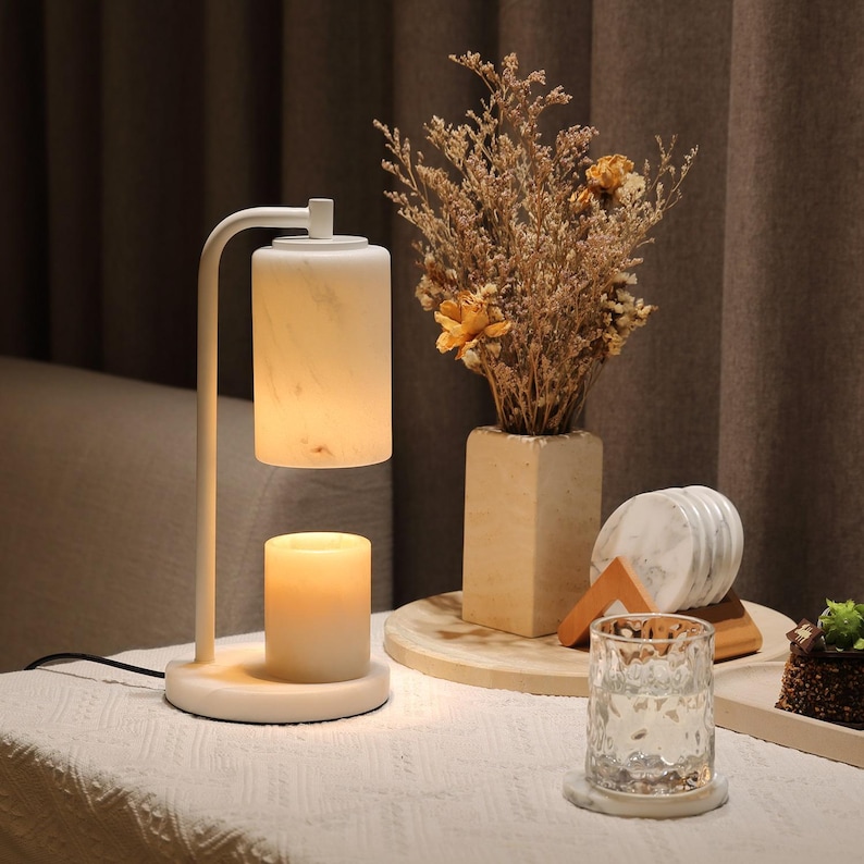 May include: A white candle warmer lamp with a cylindrical shade and base. A lit candle sits on the base. A vase with dried flowers and a glass of water with a coaster are on a table. The lamp has a curved arm.