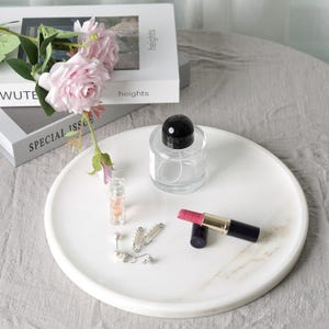 Puede incluir: Una bandeja redonda de m&aacute;rmol blanco con un frasco de perfume de vidrio transparente, un l&aacute;piz labial, aretes y un peque&ntilde;o frasco de perfume. Rosas rosas y libros en el fondo. La bandeja tiene unos 30 cm de di&aacute;metro.