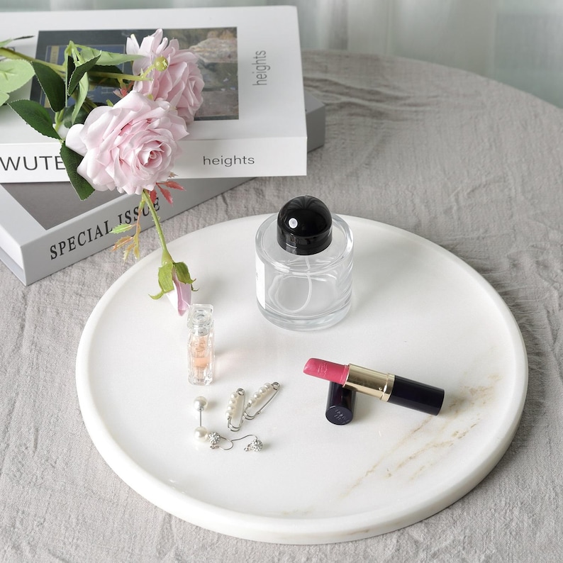 Puede incluir: Una bandeja redonda de m&aacute;rmol blanco con un frasco de perfume de vidrio transparente, un l&aacute;piz labial, aretes y un peque&ntilde;o frasco de perfume. Rosas rosas y libros en el fondo. La bandeja tiene unos 30 cm de di&aacute;metro.