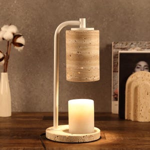 May include: A modern candle warmer lamp with a white metal frame and a cylindrical stone shade. The lamp features a white candle on a stone base. The design is minimalist and suitable for home decor.