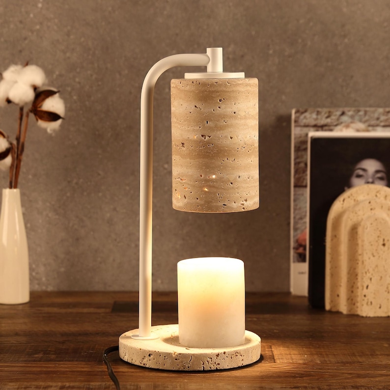 Op de afbeelding: Een kaarsverwarmerlamp met een wit metalen frame, een cilindrische kap van beige travertijnsteen en een bijpassende ronde travertijnvoet. Een witte stompkaars staat op de voet en wordt verlicht door de warmtelamp van bovenaf.