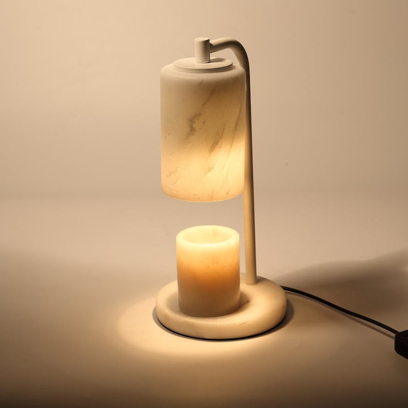 Op de afbeelding: Een kaarsverwarmerlamp van wit marmer met een cilindrische kap en een bijpassende voet. De lamp heeft een gebogen arm die de bovenste kap boven een kleiner, verlicht cilindrisch kaarsreservoir houdt en een warme gloed uitstraalt.