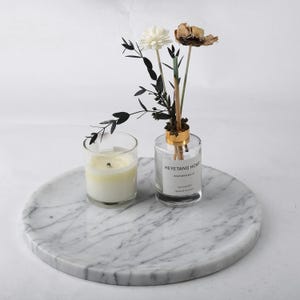 Puede incluir: Una bandeja redonda de m&aacute;rmol con una vela encendida en un vaso y un difusor de vidrio transparente con flores secas. El difusor tiene una tapa dorada y una etiqueta que dice "HEYETANG HOME". La bandeja tiene un patr&oacute;n veteado gris y blanco.