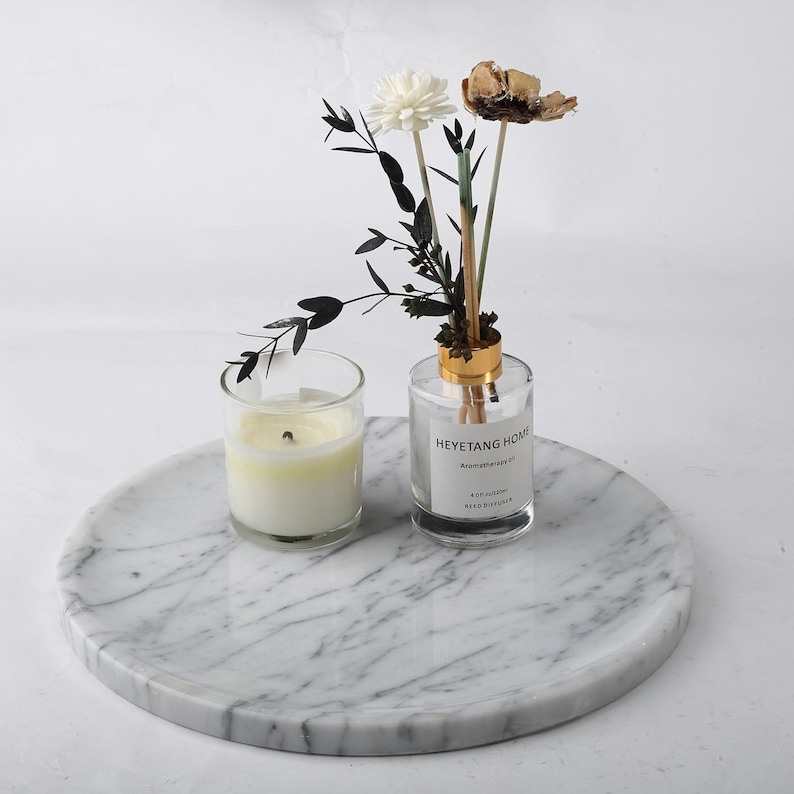 Puede incluir: Una bandeja redonda de m&aacute;rmol con una vela encendida en un vaso y un difusor de vidrio transparente con flores secas. El difusor tiene una tapa dorada y una etiqueta que dice "HEYETANG HOME". La bandeja tiene un patr&oacute;n veteado gris y blanco.