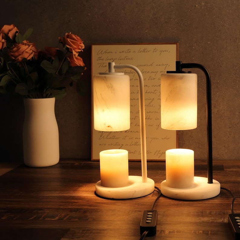 Op de afbeelding: Twee kaarsverwarmingslampen met cilindrische kappen en voetstukken met een marmerpatroon. De ene lamp heeft een witte metalen arm, de andere een zwarte metalen arm. Elke lamp verlicht een kleine, cilindrische waskaars op de ronde marmeren voet.