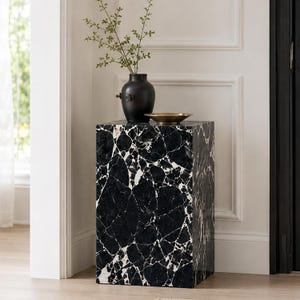 May include: A rectangular pedestal table featuring a black and white marble pattern with large, angular veining. On top of the pedestal sits a matte black ceramic vase containing a green leafy branch and a small, shallow metallic gold bowl.