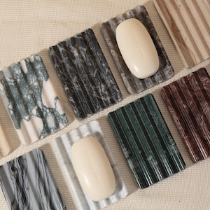 May include: A collection of soap dishes and soaps. The soap dishes are made of stone with various patterns and colors, including white, gray, black, and green. The soaps are oval-shaped and light-colored.