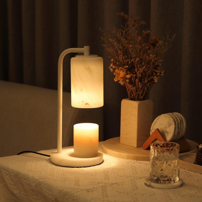 Op de afbeelding: Een witte kaarsverwarmerlamp met een cilindervormige kap met marmerpatroon en een bijpassende ronde voet. De lamp verlicht een witte stompkaars die direct onder de kap staat en verspreidt een warme, zachte gloed over de omgeving.