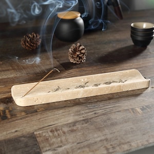 May include: A beige stone incense holder with a burning incense stick, releasing smoke. The holder is on a wooden surface, with pine cones and a black ceramic pot in the background. Small black bowls are also visible.