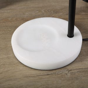 Op de afbeelding: Een ronde witte marmeren voet met een glad, gepolijst oppervlak en subtiele grijze aders. Een enkele matzwarte metalen stang steekt verticaal uit de zijkant van de voet en verbindt zich met een structuur die buiten het beeld valt.