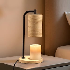 Op de afbeelding: Een kaarsverwarmerlamp met een matzwart metalen frame, een cilindrische beige travertijn stenen kap en een bijpassende ronde travertijn voet. Een witte waskaars staat op de voet en wordt verlicht door de warmte van de lamp erboven.