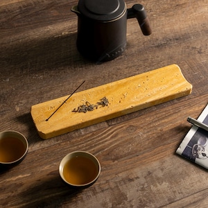May include: A tea ceremony setup. A rectangular, light-yellow stone tray holds tea leaves and an incense stick. A black teapot with a wooden handle is above the tray. Two teacups and a newspaper are on the wooden surface.
