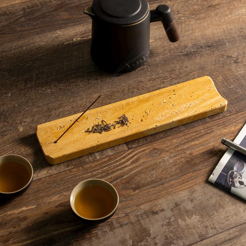 May include: A tea ceremony setup. A rectangular, light-yellow stone tray holds tea leaves and an incense stick. A black teapot with a wooden handle is above the tray. Two teacups and a newspaper are on the wooden surface.