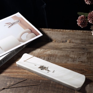 May include: A white marble incense holder, rectangular with curved edges. It features a small hole for the incense stick and a small pile of ash. The holder sits on a wooden surface, with a book and flowers in the background.