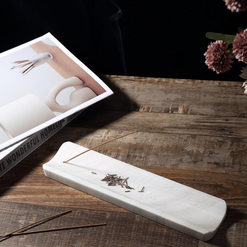 May include: A white marble incense holder, rectangular with curved edges. It features a small hole for the incense stick and a small pile of ash. The holder sits on a wooden surface, with a book and flowers in the background.