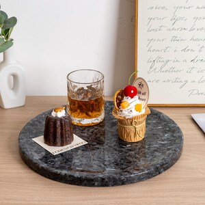 Puede incluir: Una bandeja redonda de m&aacute;rmol gris oscuro con un vaso de l&iacute;quido &aacute;mbar, un peque&ntilde;o pastel de chocolate y una magdalena con una cereza encima. La bandeja est&aacute; sobre una superficie de madera, con un poema enmarcado en el fondo.