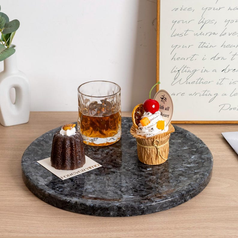 Puede incluir: Una bandeja redonda de m&aacute;rmol gris oscuro con un vaso de l&iacute;quido &aacute;mbar, un peque&ntilde;o pastel de chocolate y una magdalena con una cereza encima. La bandeja est&aacute; sobre una superficie de madera, con un poema enmarcado en el fondo.