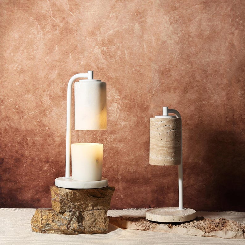 May include: Two candle warmers with white metal frames and cylindrical stone shades. The left warmer features a smooth white marble shade and base, while the right warmer has a textured beige travertine shade and base, both emitting light.