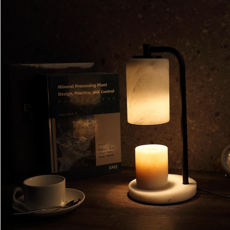 Op de afbeelding: Een kaarsverwarmerlamp met een witte marmeren voet en cilindrische kap, voorzien van een zwarte metalen arm. Een brandende kaars staat op de voet en verspreidt een warme gloed. Daarnaast een boek met de titel Mineral Processing Plant Design.