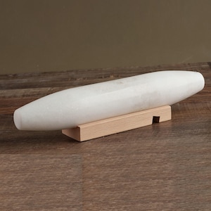 May include: A smooth, white, cylindrical stone object with tapered ends resting horizontally on a light-colored wooden stand. The stone has a polished finish and a subtle marbled texture, while the wooden base features a rectangular notch on one side.