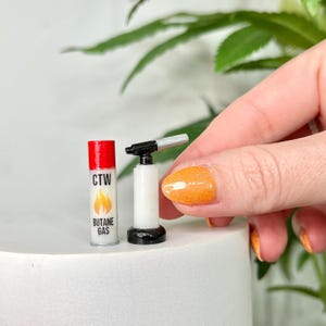May include: Miniature butane torch and a red and white butane gas canister. The canister has the text "CTW" and "BUTANE GAS" with a flame graphic. The torch is white with a black handle and silver nozzle.